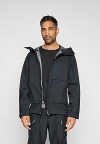 Giacca impermeabile nera con cappuccio, chiusura frontale con cerniera e dettagli grigi. Presenta molteplici tasche e un design a vestibilità comoda.