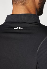 Black athletic polo shirt featuring a textured collar, smooth fabric, and a small white logo on the back near the neck.