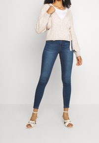 Žena v béžovém svetr s překládaným designem, bílém tričku, tmavě modrých skinny džínách, bílých sandálech na podpatku a nese béžovou kabelku přes rameno.