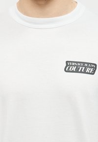 Λευκό μπλουζάκι με στρογγυλή λαιμόκοψη και το λογότυπο "VERSACE JEANS COUTURE" σε μαύρο και άσπρο στην αριστερή πλευρά του στήθους.