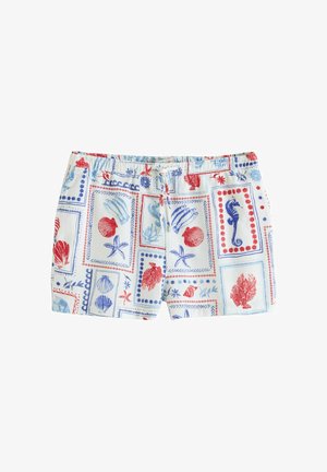 Femeie purtând cămașă și pantaloni scurți cu imprimeu cu scoici, stând desculță pe malul stâncos cu oceanul în fundal, uitându-se lateral.