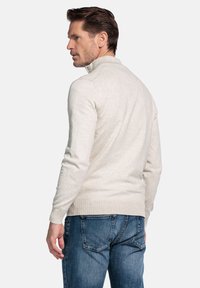 Beige gebreide trui met een hoge kraag, geribbelde manchetten en zoom. Draagt blauw spijkerbroek. Heeft een aansluitend ontwerp en een gladde textuur.