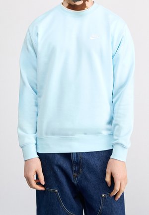 Osoba ubrana w jasnoniebieską bluzę Nike w połączeniu z ciemnoniebieskimi dżinsami, na jednolitym jasnym tle.
