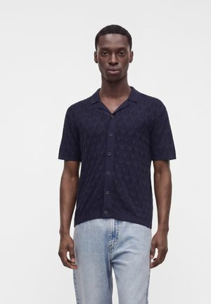 Homme portant une chemise bleu marine à manches courtes boutonnée avec un motif texturé et un jean bleu clair, debout devant un fond uni.
