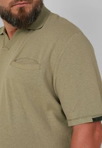 Olijfgroene geribbelde poloshirt met een korte placket en een klein borstzakje. Heeft een gestructureerd oppervlak en een logolabel op de mouw.