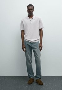 Polo grigio chiaro a maniche corte, paio di jeans azzurri chiari e scarpe in suede marrone, in piedi su un pavimento scuro e texture.