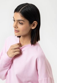 Jasnoróżowy sweter z okrągłym dekoltem, o miękkiej fakturze i opuszczonych ramionach. Modelka dostosowuje dekolt ręką.