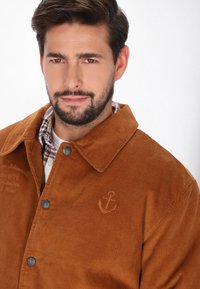 Brun kordfløjl jakke med krave, sølvknapper og detaljer med broderet anker. Båret over en ternet skjorte.