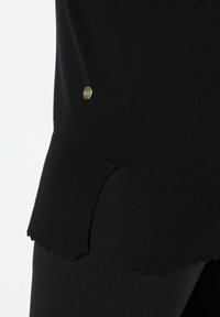 Musta neulepaita, jossa on sakaroitettu helma ja kultainen metallinen logoakseli sivussa. Pehmeä rakenne ja istuva muotoilu.