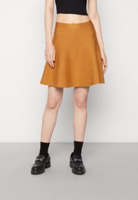 Une jupe A-line moutarde en tissu texturé, avec une taille haute et une finition lisse, associée à des mocassins noirs et des chaussettes.