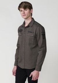 Langermige, knoopsluiting overhemd van donkergrijze stof, voorzien van twee voorzakken, geborduurde patches op de schouders en een kraag.