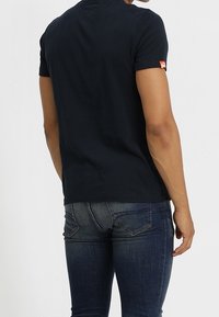 Camiseta corta azul marino de algodón, con cuello redondo y una pequeña etiqueta roja en la mangas izquierda. Combinada con jeans ajustados.
