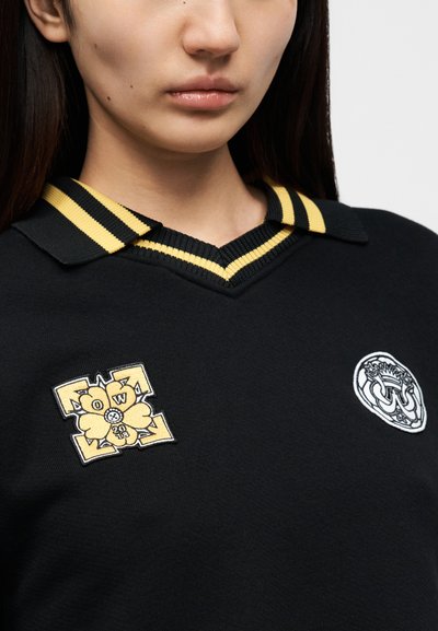 Μαύρο πόλο μπλουζάκι με κολάρο με κίτρινες και μαύρες ρίγες. Διαθέτει δύο κεντημένα εμβλήματα: ένα με σχέδιο λουλουδιού και ένα κυκλικό έμβλημα.