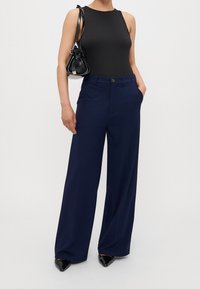 Svart ärmlös bodysuit, marinblå vida byxor med knappstängning, fickor och svart handväska, kombinerat med svarta pumps med spetsig tå.