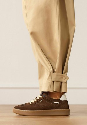 Bruine suède sneaker met witte accenten gedragen met beige broek met een met knopen afgeremde band bij de enkel, staand op een houten vloer.