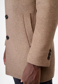 Bruin wollen jas met een gestructureerde afwerking, voorzien van een dubbellaags ontwerp, zwarte knopen en een zijzakdetail aan de linkerkant.