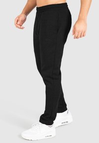 Schwarze Baumwoll-Jogginghosen mit elastischem Bund, schmalem Schnitt und dezentem Logo-Detail in der Nähe der Tasche. Getragen mit weißen Turnschuhen.