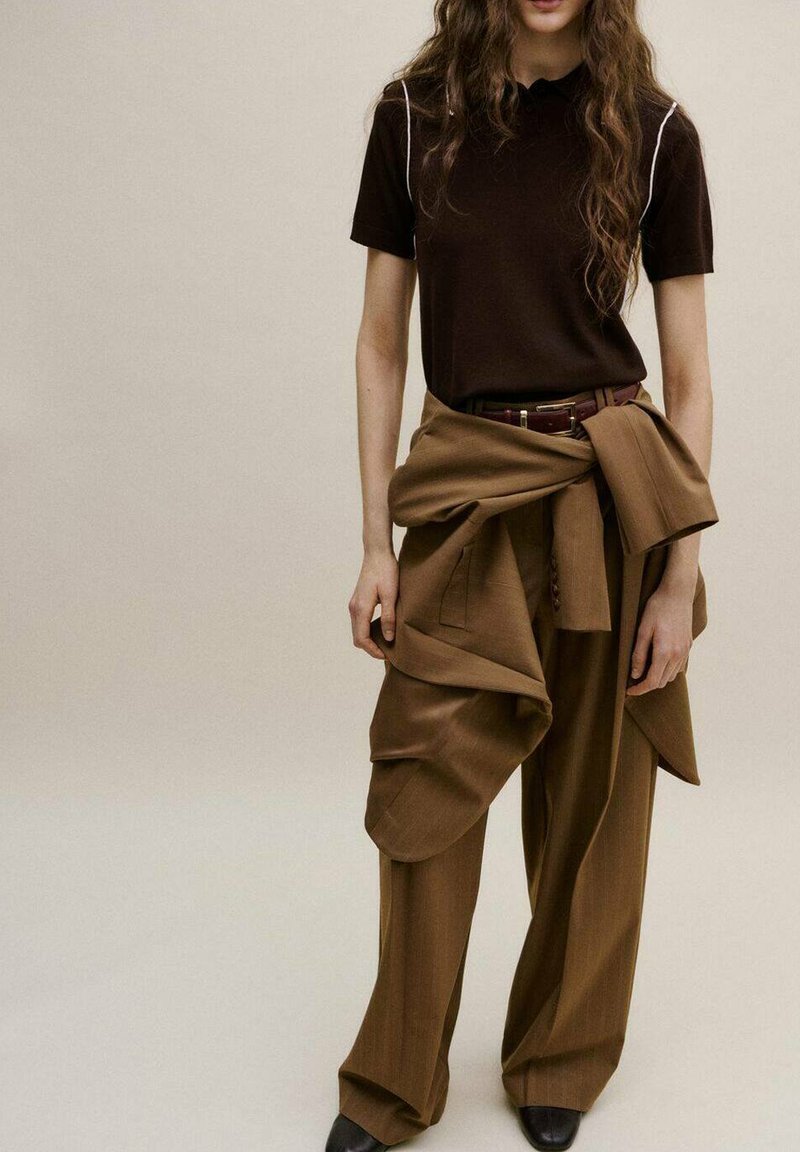 Woman wearing brown wide-leg pants, a matching brown jacket tied around the waist, and a dark brown short-sleeve top with white piping.