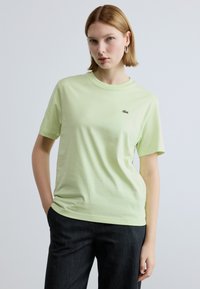 Ανοιχτό πράσινο βαμβακερό t-shirt με στρογγυλή λαιμόκοψη, κοντά μανίκια και μικρό κεντημένο λογότυπο στο στήθος. Φορεμένο με σκούρο τζιν.