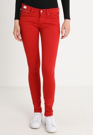 Obcisłe, jasnoczerwone dżinsy noszone z białymi trampkami i czarną bluzką z długim rękawem, pokazane od talii do stóp na białym tle.