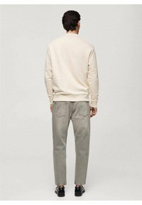 Mężczyzna w kremowej bluzie, szarych dżinsach i czarnych butach, stojący tyłem do kamery na jednolitym jasnym tle.