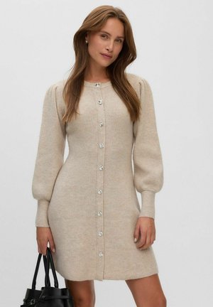 Mujer con cabello largo y castaño, vestida con un vestido de punto beige con botones y mangas abullonadas, sosteniendo un bolso negro sobre un fondo liso.