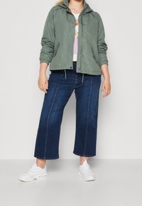 Light green, zip-up jacket with a hood, cinched waist, worn over a white top and dark blue wide-leg jeans. Footwear is white sneakers.