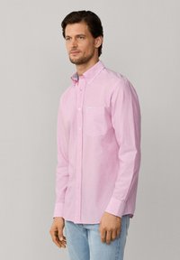 Man draagt een lichtroze overhemd met knopen en een borstzak, gecombineerd met lichtblauwe jeans, staande tegen een effen achtergrond.