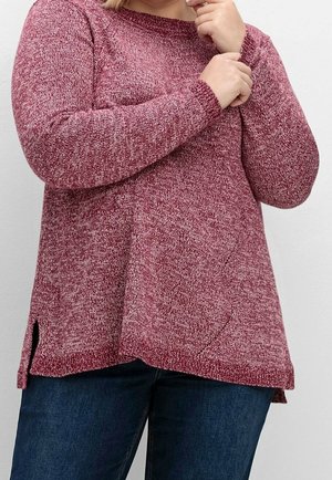 Osoba ubrana w bordowy sweter z długim rękawem z dzianiny z rozcięciami po bokach, w połączeniu z ciemnoniebieskimi dżinsami, z rękami złożonymi przy twarzy.