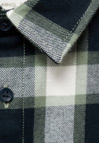 Vihreä ja musta ruudullinen paidankaulus, jossa on napitusyksityiskohta, ja siinä on teksturoitu kudonta sekä kontrastina valkoiset neliöt ruutukuviossa.