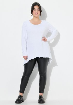 Weißes Langarmoberteil mit rundem Ausschnitt und asymmetrischem Saum, kombiniert mit schwarzen Fauxleder-Leggings und klobigen schwarzen Schuhen.