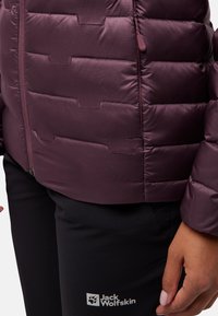 Giacca trapuntata bordeaux con finitura liscia, caratterizzata da una zip frontale e un dettaglio con logo. Abbinata a pantaloni neri.