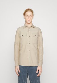 Beige long-sleeve shirt made of woven fabric, featuring two chest pockets, black buttons, and a pointed collar. Smooth texture.