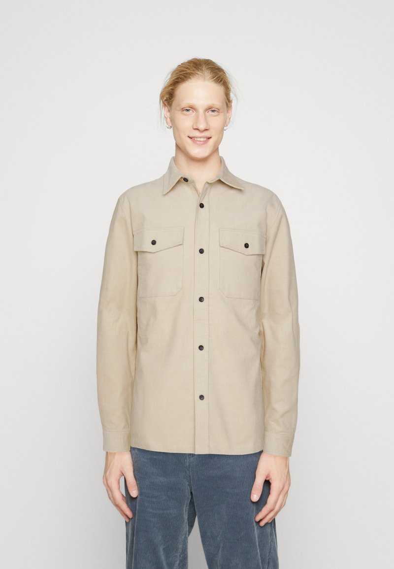 Beige long-sleeve shirt made of woven fabric, featuring two chest pockets, black buttons, and a pointed collar. Smooth texture.