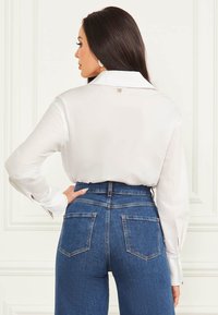 Blusa di satin bianco con colletto a punta, dotata di un piccolo logo sul retro; abbinata a jeans in denim blu a vita alta con tasche posteriori.