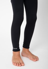 Schwarze Leggings aus dehnbarem Material, mit gepolsterten Knieflicken und einem Logo nahe dem Saum. Glatte Textur und figurbetontes Design.