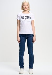 Bílý tričko s krátkým rukávem a grafikou "BIG STAR" ve tmavém písmu. Odpovídající tmavě modré džínsy a bílé tenisky. Jednoduchý, přiléhavý design.