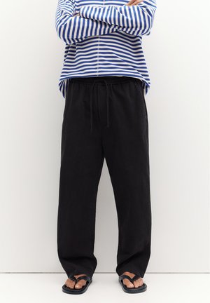 Personne portant un pantalon noir ample avec taille à cordon, une chemise à manches longues rayée bleu et blanc, et des tongs noires, debout contre un mur blanc.