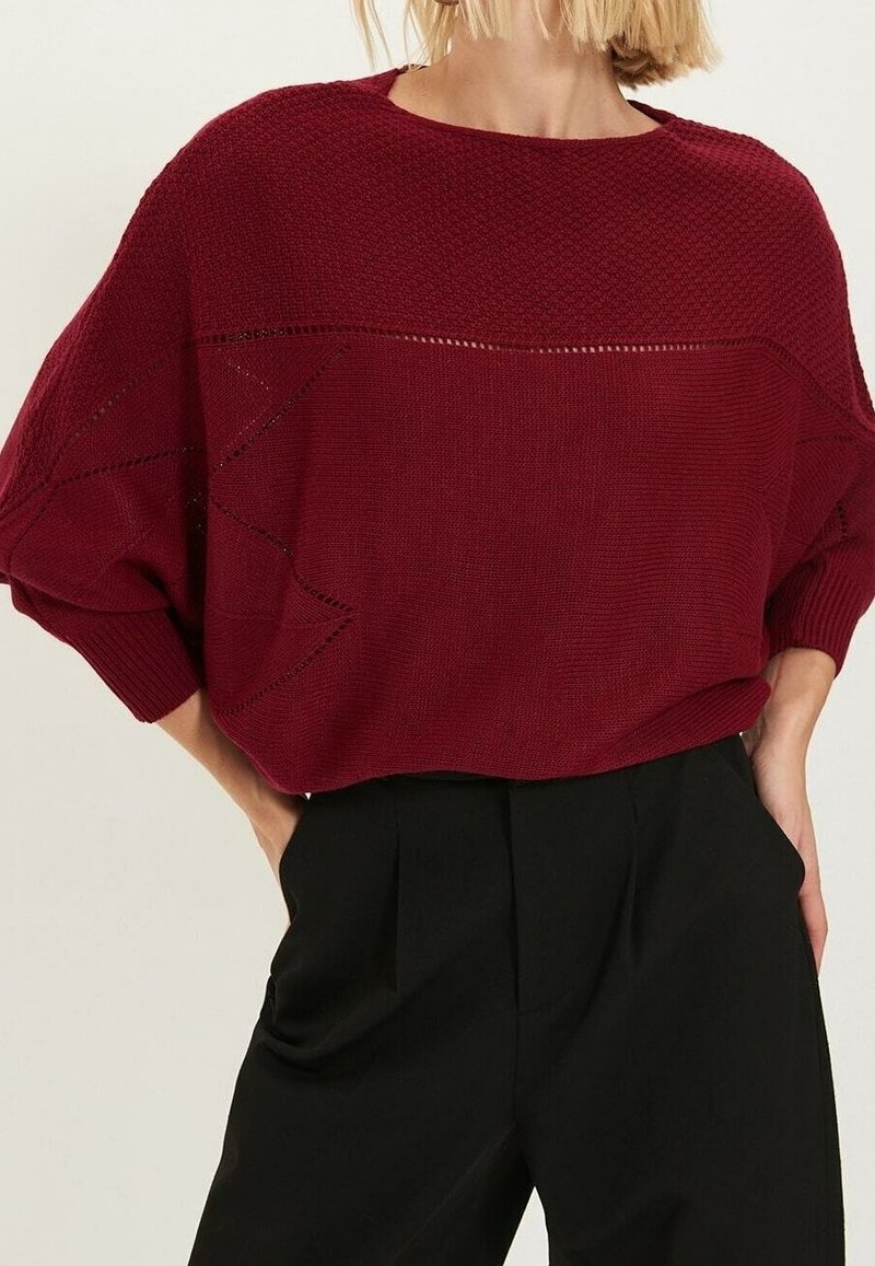Pull en maille bordeaux oversize avec une section supérieure texturée, doté d'un large col et de détails en motif ajouré. Associé à un pantalon noir.