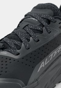 Zwarte sportieve schoen met een bovenwerk van mesh, textuur overlays en reflecterende accenten. Voorzien van platte veters en een gevoerde zool voor comfort.