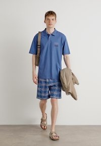 Blå piké med en liten grafisk patch, rutiga denimshorts, beige jacka i handen och bruna sandaler med öppen tå.