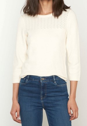 Licht crèmekleurige gebreide trui met driekwart mouwen, een gestructureerd lijfje en een patroon op het beleg, gecombineerd met donkerblauwe, hooggetailleerde jeans.