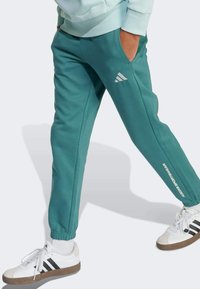 Tyrkysové teplákové nohavice s elastickým pásom, bočnými vreckami a bielym nápisom "ADIDAS SPORTWEAR". Skombinované s bielymi teniskami s čiernymi pruhmi.