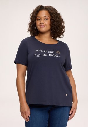 Kobieta o kręconych włosach, ubrana w granatowy t-shirt z włoskim tekstem oraz rysunkami słońca i chmur, w połączeniu z niebieskimi dżinsami na jednolitym tle.