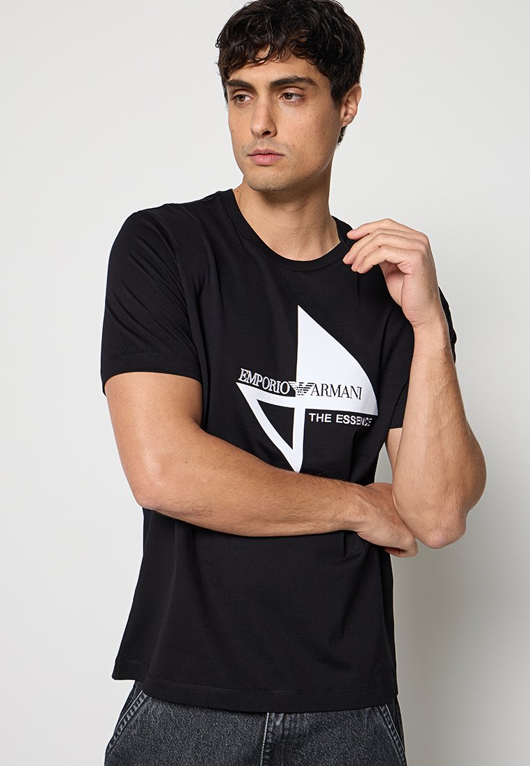 Joven con camiseta negra Emporio Armani con logo geométrico blanco y texto, vistiendo vaqueros oscuros, posando frente a un fondo claro y liso.