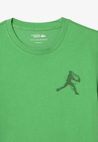 Vihreä puuvillapaita pyöreällä kauluksella, säännöllinen istuvuus, jossa tumma siluetti pelaajasta ja hienovarainen teksti kauluksessa.