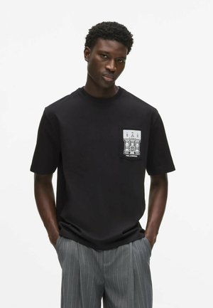 Homme portant une chemise noire à manches courtes avec une poche blanche graphique et un pantalon gris à fines rayures, debout les mains dans les poches.