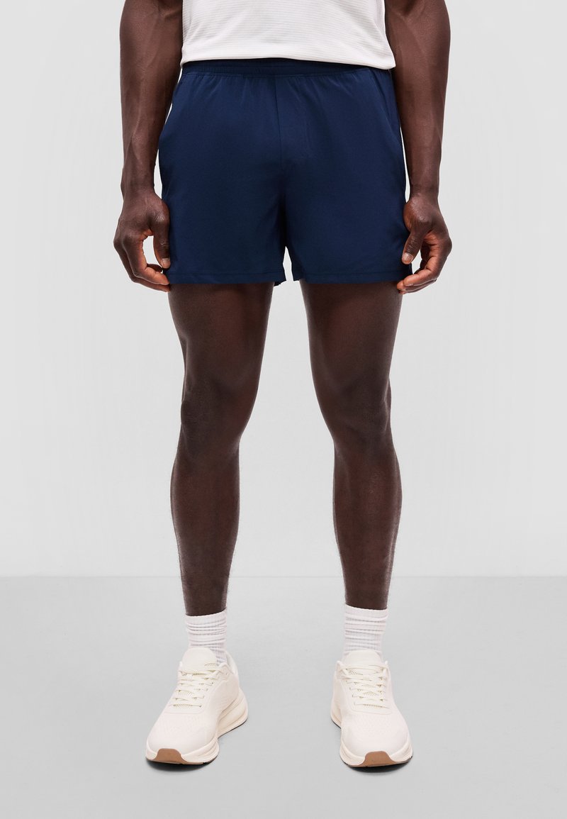 Uomo che indossa pantaloncini sportivi blu navy, calzini bianchi e scarpe da corsa bianche, in piedi su uno sfondo grigio uniforme.
