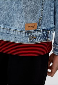 Chaqueta de denim azul claro con una etiqueta marrón de Pull&Bear sobre una camiseta de rayas rojas y negras, llevada por una persona con una mano visible.