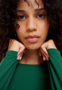 Zielona bluzka z długim rękawem z miękkiego materiału, z szerokim dekoltem. Ręce modelki spoczywają na jej ramionach, podkreślając gładką skórę i lśniące usta.